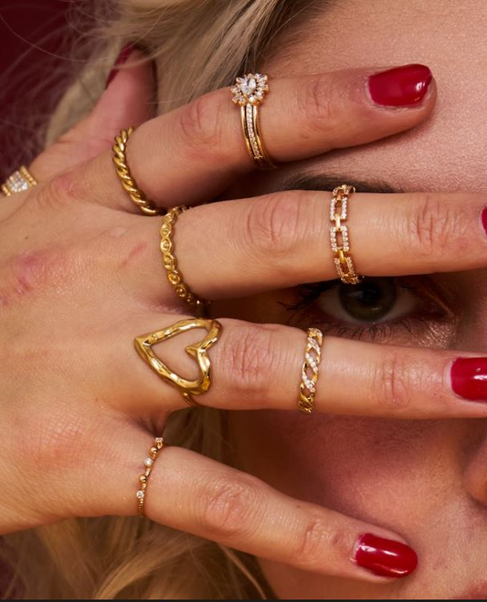 Hand mit mehreren goldfarbenen Ringen, darunter ein herzförmiger und ein gedrehter Ring, rote Fingernägel