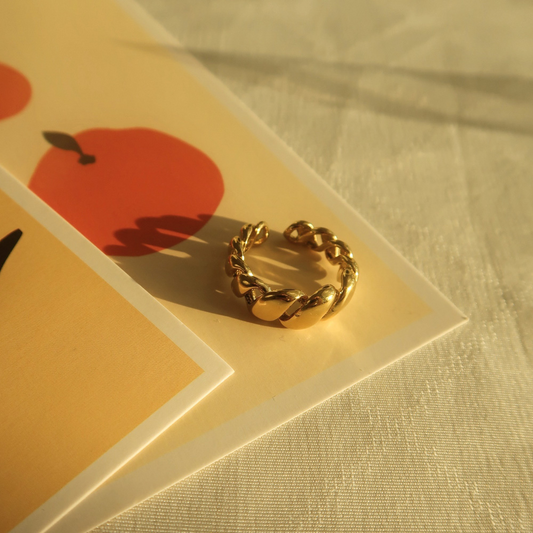 Goldfarbener Edelstahlring mit gedrehter Struktur auf beigem Hintergrund mit rotem Apfelmotiv