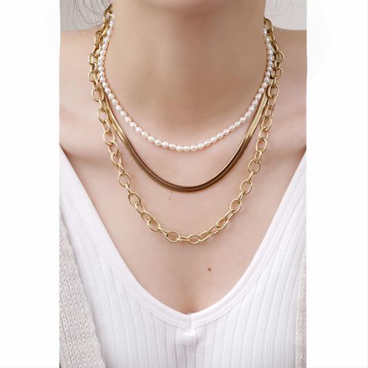 Drei Halsketten übereinander: goldene Gliederkette, goldene Schlangenkette, Perlenkette auf weißem Oberteil.