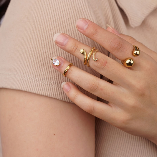 Hand mit mehreren goldfarbenen Ringen, darunter ein glänzender Edelstahlring, auf beigem Hintergrund.