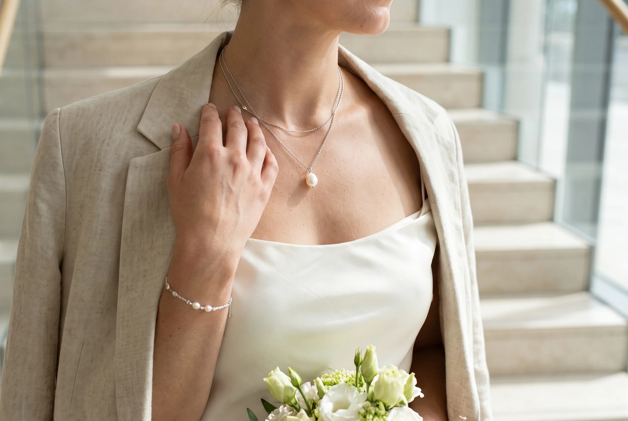 Braut vor einem modernen Standesamt in schlichtem elfenbeinfarbenem Look mit feiner Süßwasserperlen-Kette und zartem Perlenarmband.