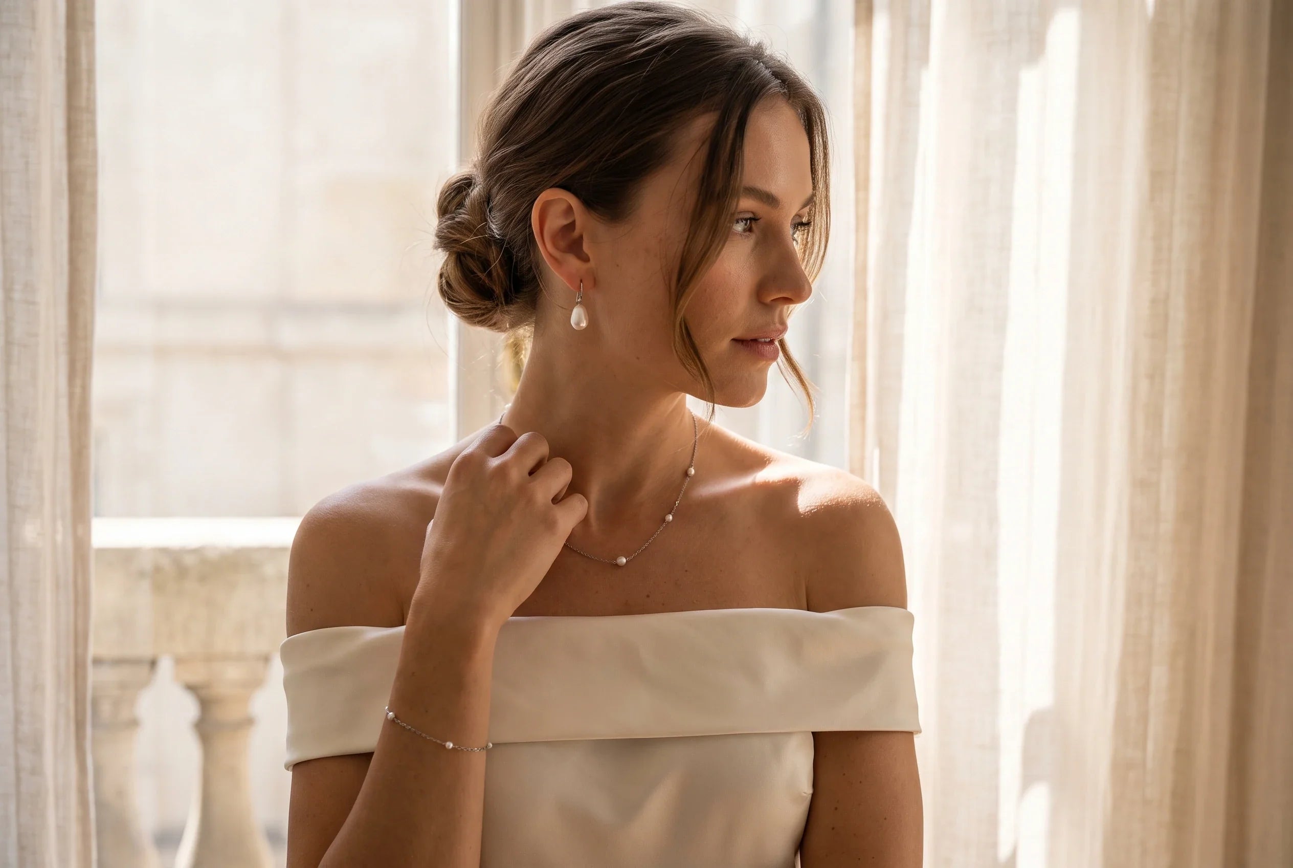 Person mit lockerem tiefem Chignon und schulterfreiem hellen Kleid trägt sichtbare Perlenhänger; dazu eine feine Kette und ein zartes Armband.