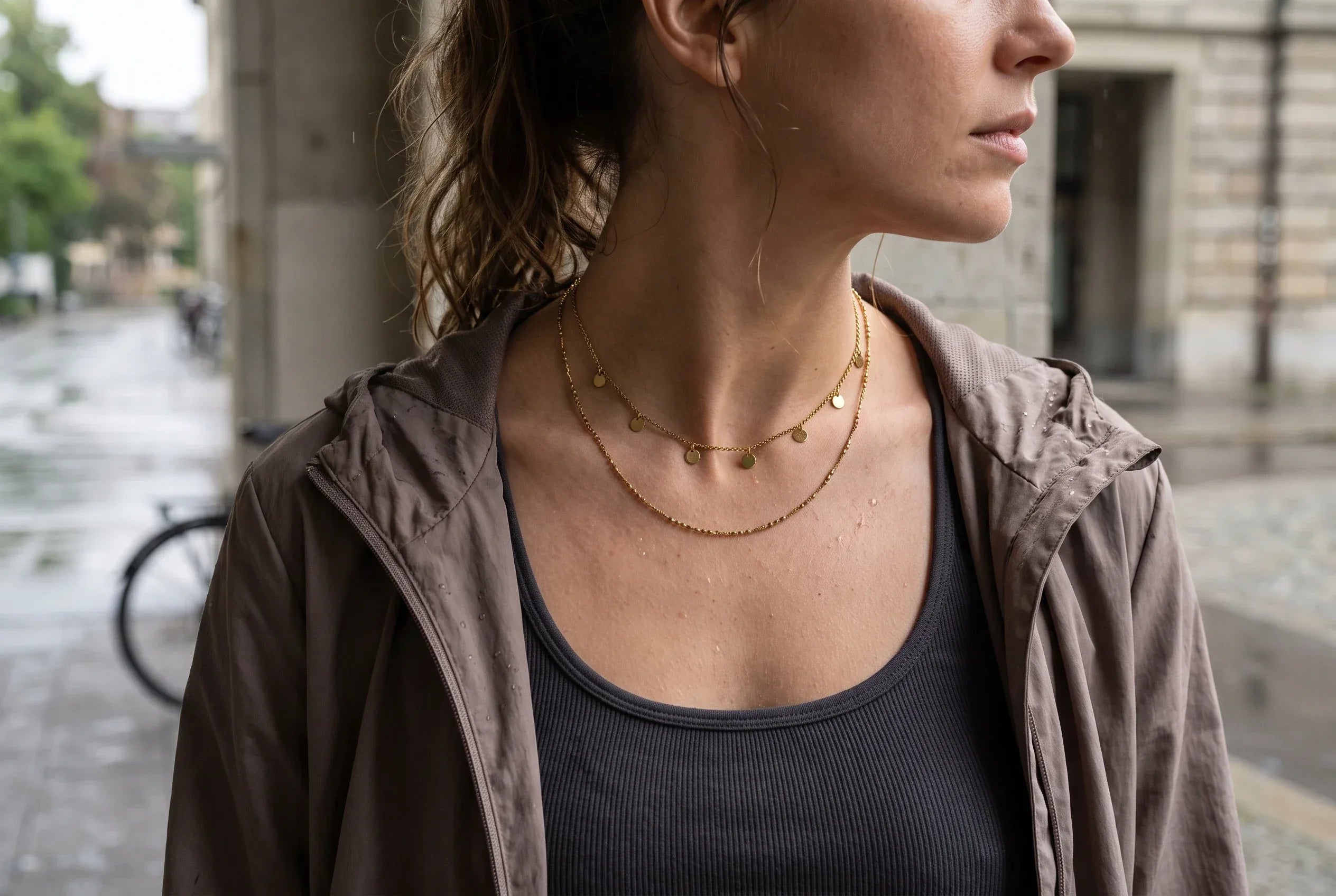 Person nach Workout und Sommerregen in grauem Tanktop und offenem Windbreaker; am Dekolleté liegen zwei feine goldfarbene Edelstahlketten in unterschiedlichen Längen.