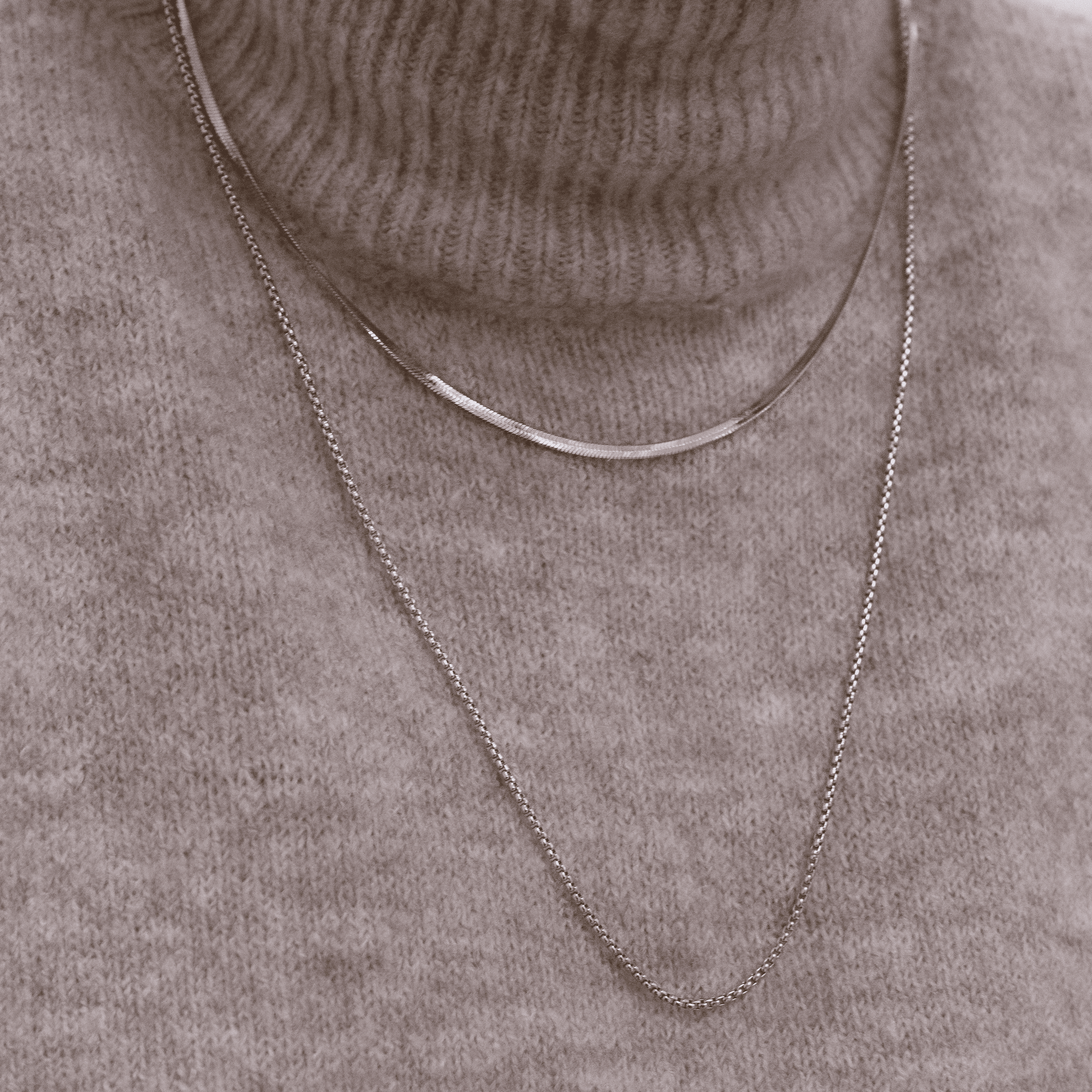 Zwei Edelstahlketten in Schlangenoptik, eine kürzer und eine länger, auf grauem Strickpullover drapiert.